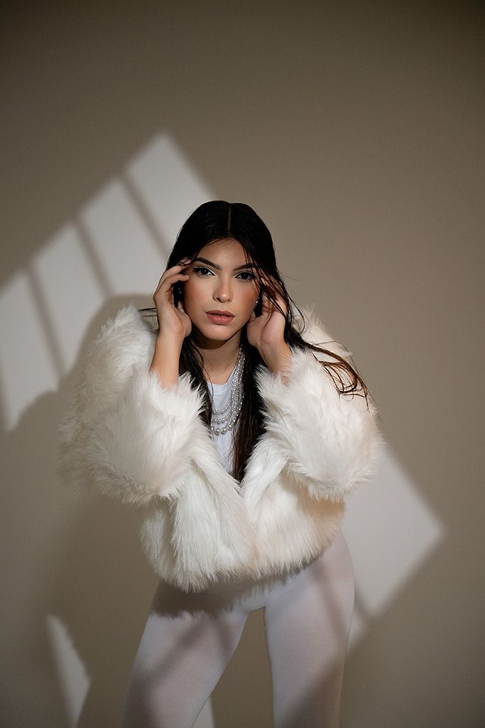 Woman in white fluffy fur coat and white pants posing with hands near face in soft natural light.