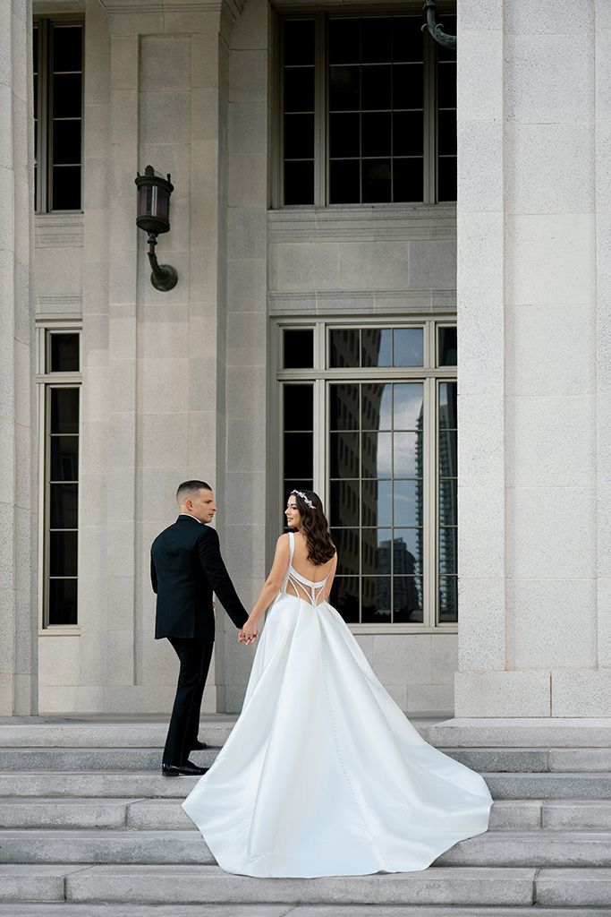 Bride and groom holding hands on steps of grand stone building, bride in flowing white gown.