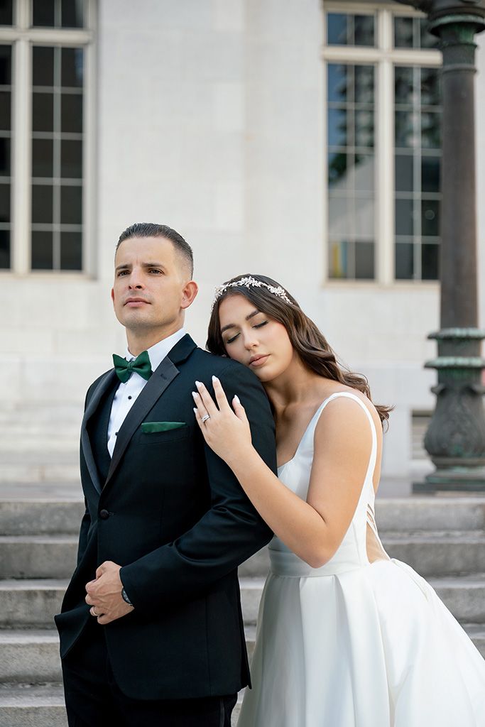 Bride in white gown leans on groom in black tuxedo with green bow tie on elegant stone steps.