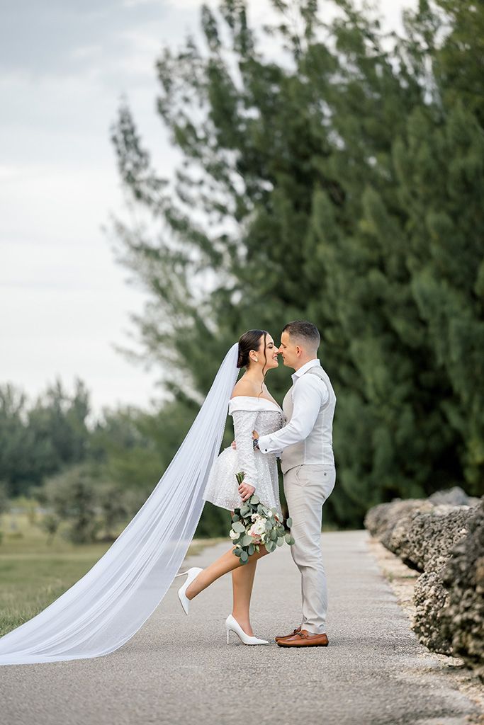 Bride and groom share a romantic kiss on a tree-lined path, bride wearing white off-shoulder dress with long veil.