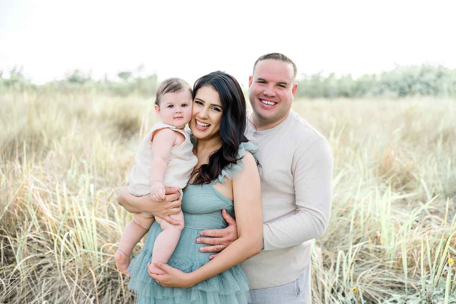 A smiling family of three poses in a golden field, with a pregnant mother in a teal dress holding a baby.