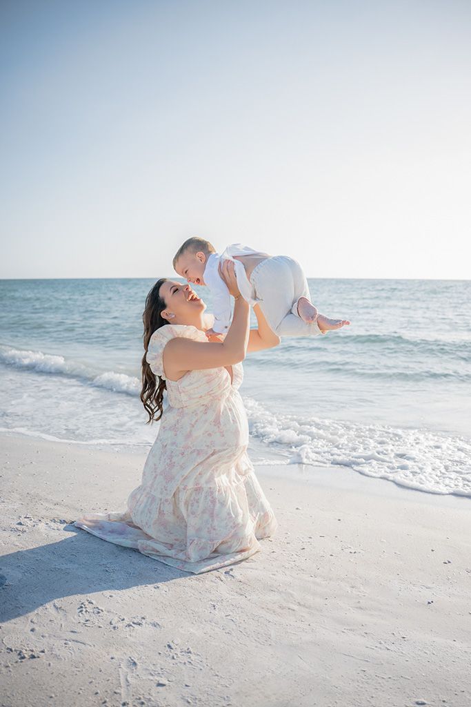 Mother in floral dress kneeling on sandy beach, joyfully lifting her baby up in the air by the ocean.