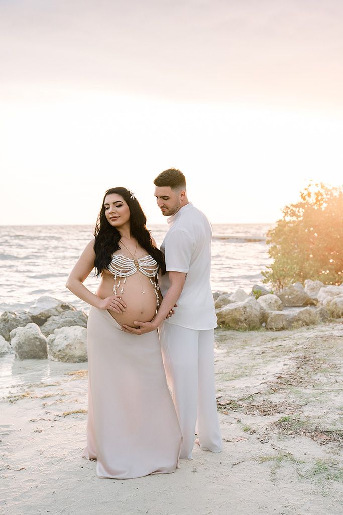 Couple embracing pregnant belly in elegant attire on a sandy beach at golden sunset during maternity shoot.
