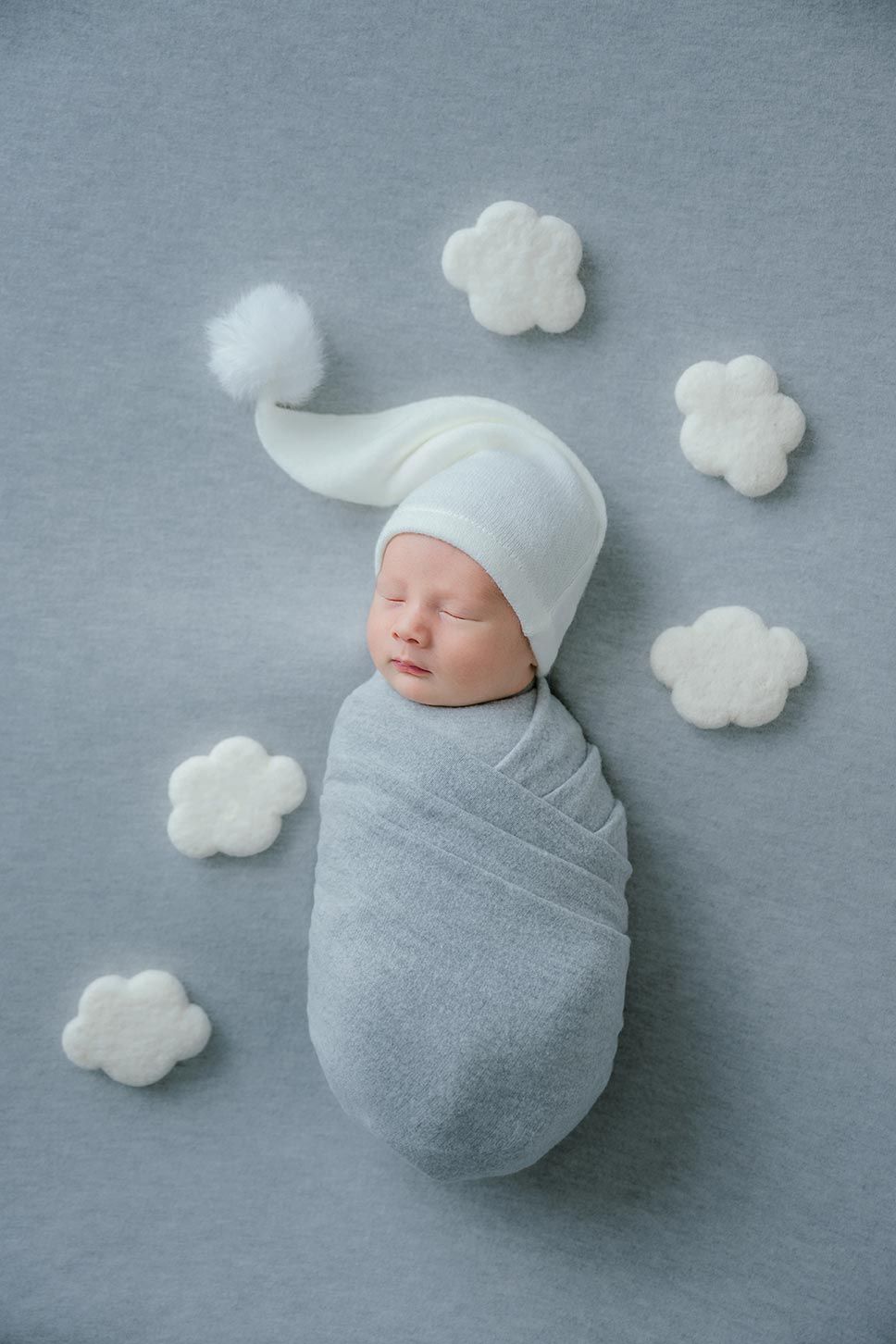 Peaceful newborn baby swaddled in gray wrap wearing white knit sleep cap surrounded by cloud decorations.