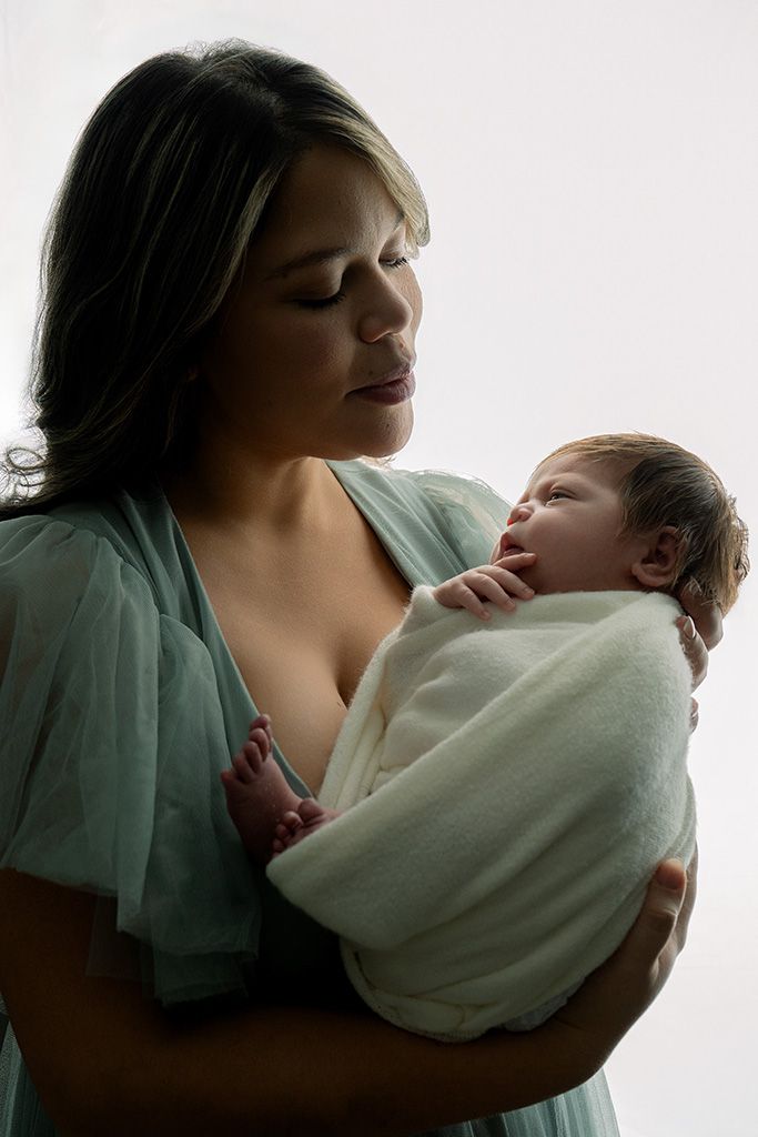 A mother tenderly gazes at her swaddled newborn baby wrapped in white, both bathed in soft light.