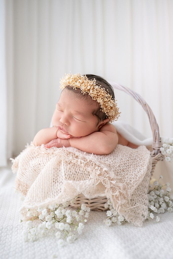 Sleeping newborn baby wearing a gold floral crown resting in a wicker basket with cream lace and white flowers.