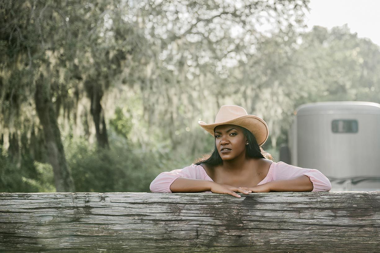 Person wearing a brown hat leans against a rustic wooden fence in a dreamy Spanish moss-draped Southern landscape.