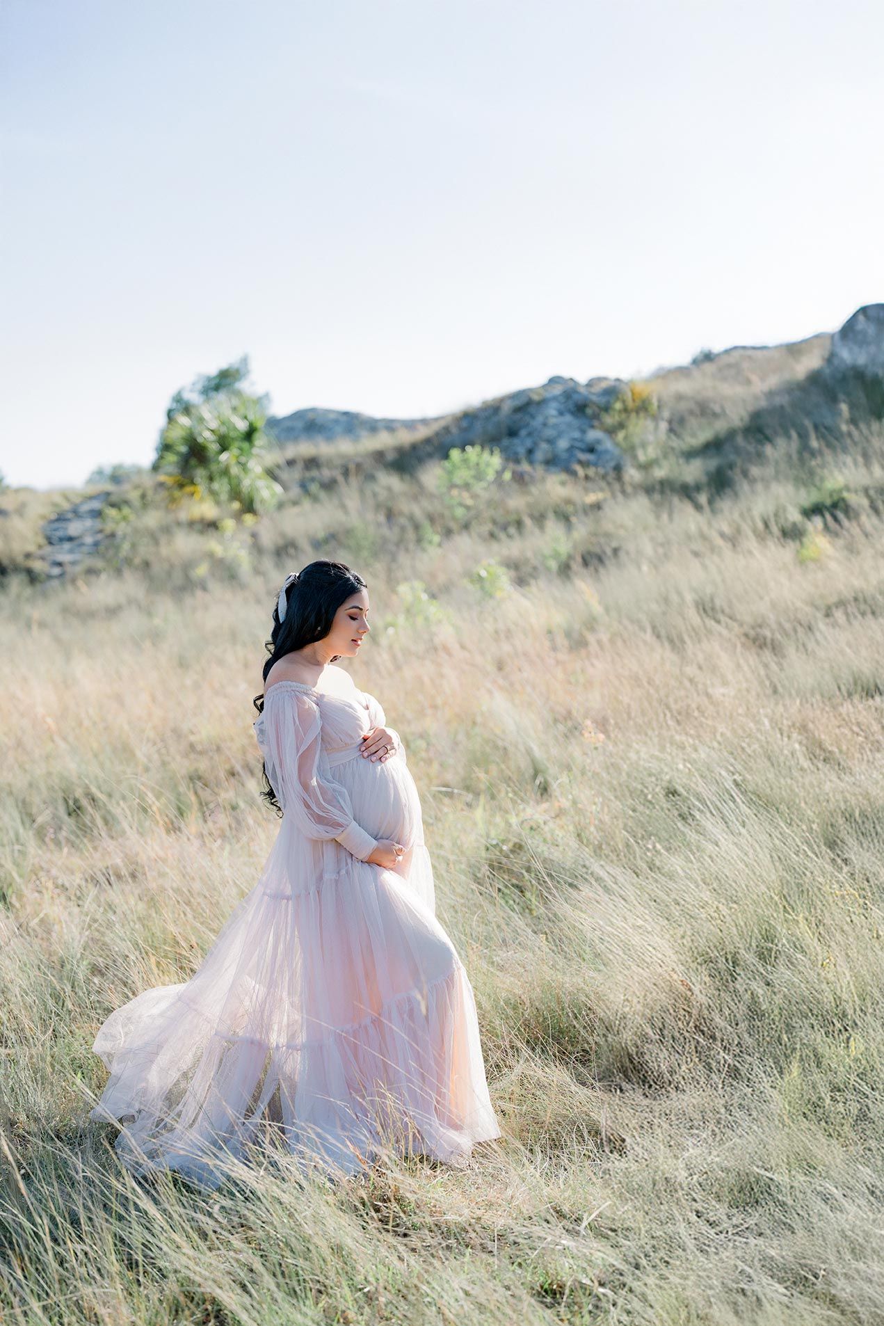 Flowing pink maternity gown billows in the wind against a scenic backdrop of golden hillside grasses and mountains.