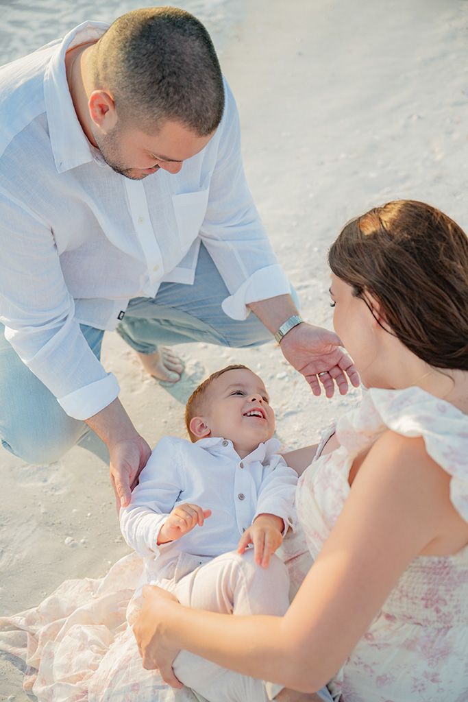 Parents playing with laughing baby on sandy beach in warm sunlight.