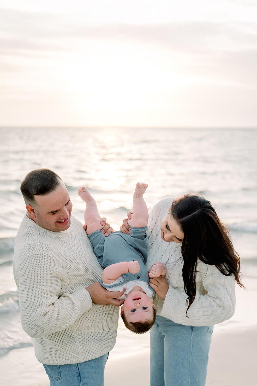 Family of three enjoying a beach sunset, parents playfully holding baby upside down, wearing matching white sweaters and jeans.