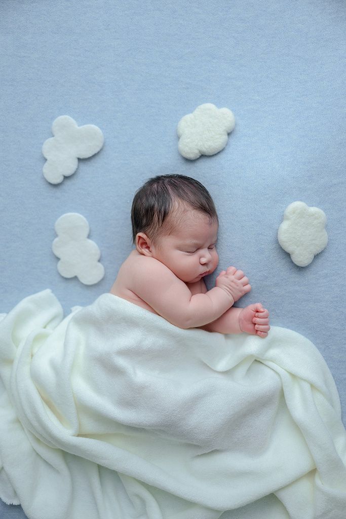 Sleeping newborn baby wrapped in white blanket on blue background with white felt clouds.