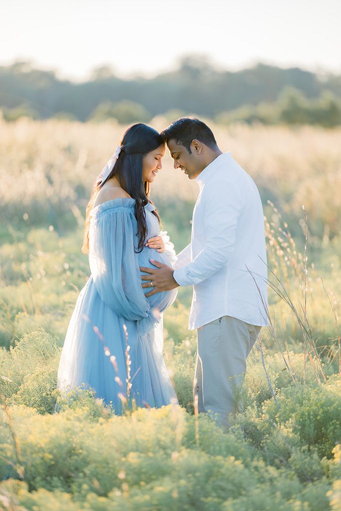 Couple in a sunlit field, woman in blue maternity gown, man in white, foreheads touching, hands on baby bump.