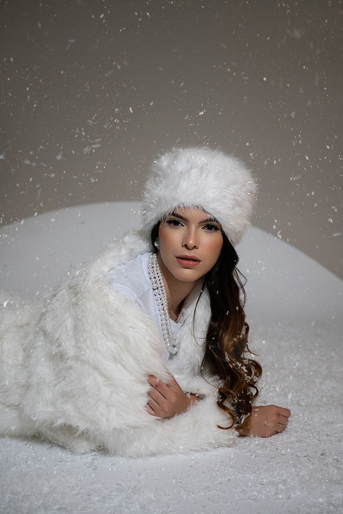 Woman in white faux fur coat and hat lies in snow with snowflakes falling around her.