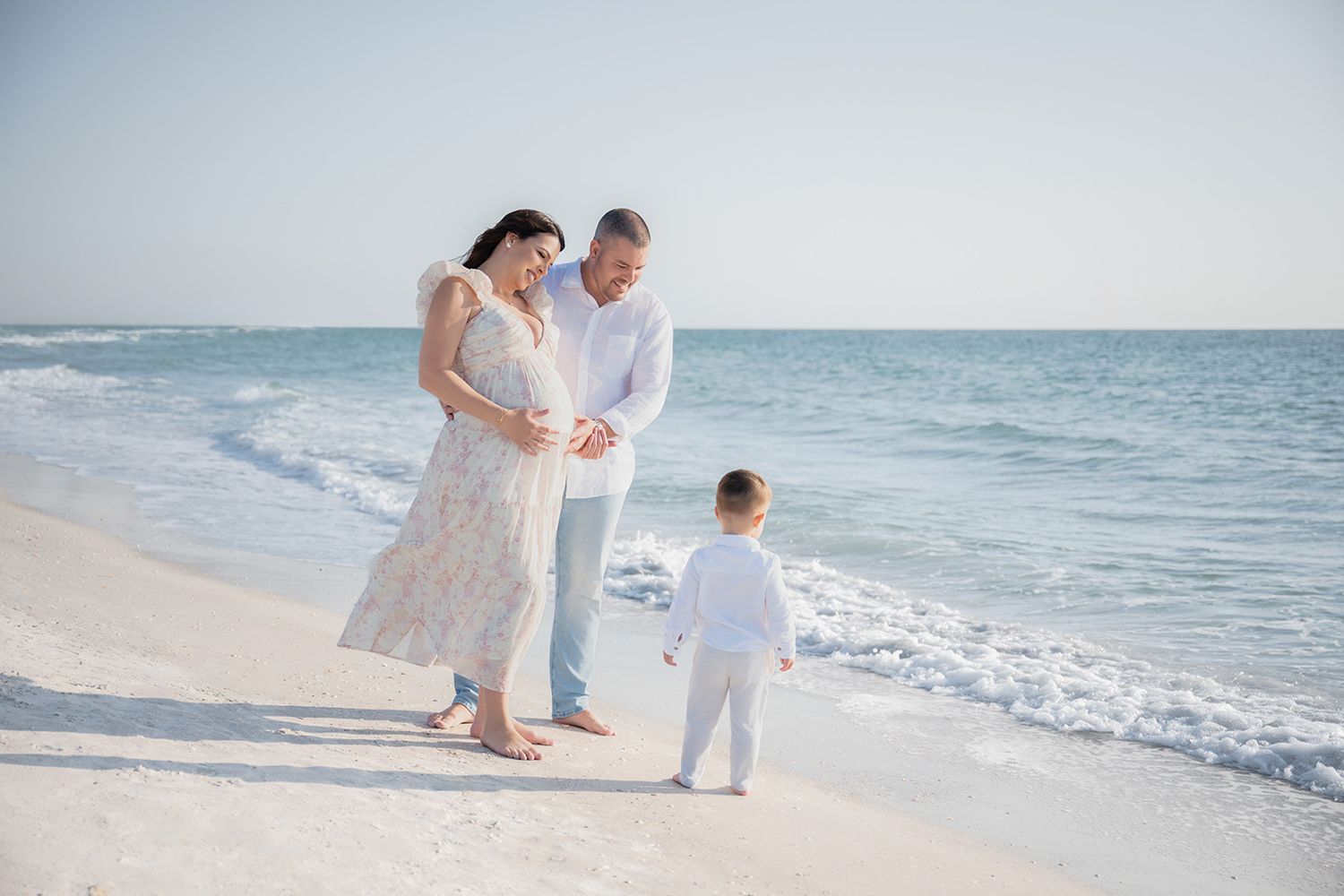 Pregnant woman and man admire toddler on sunny white sand beach with calm turquoise ocean waves.