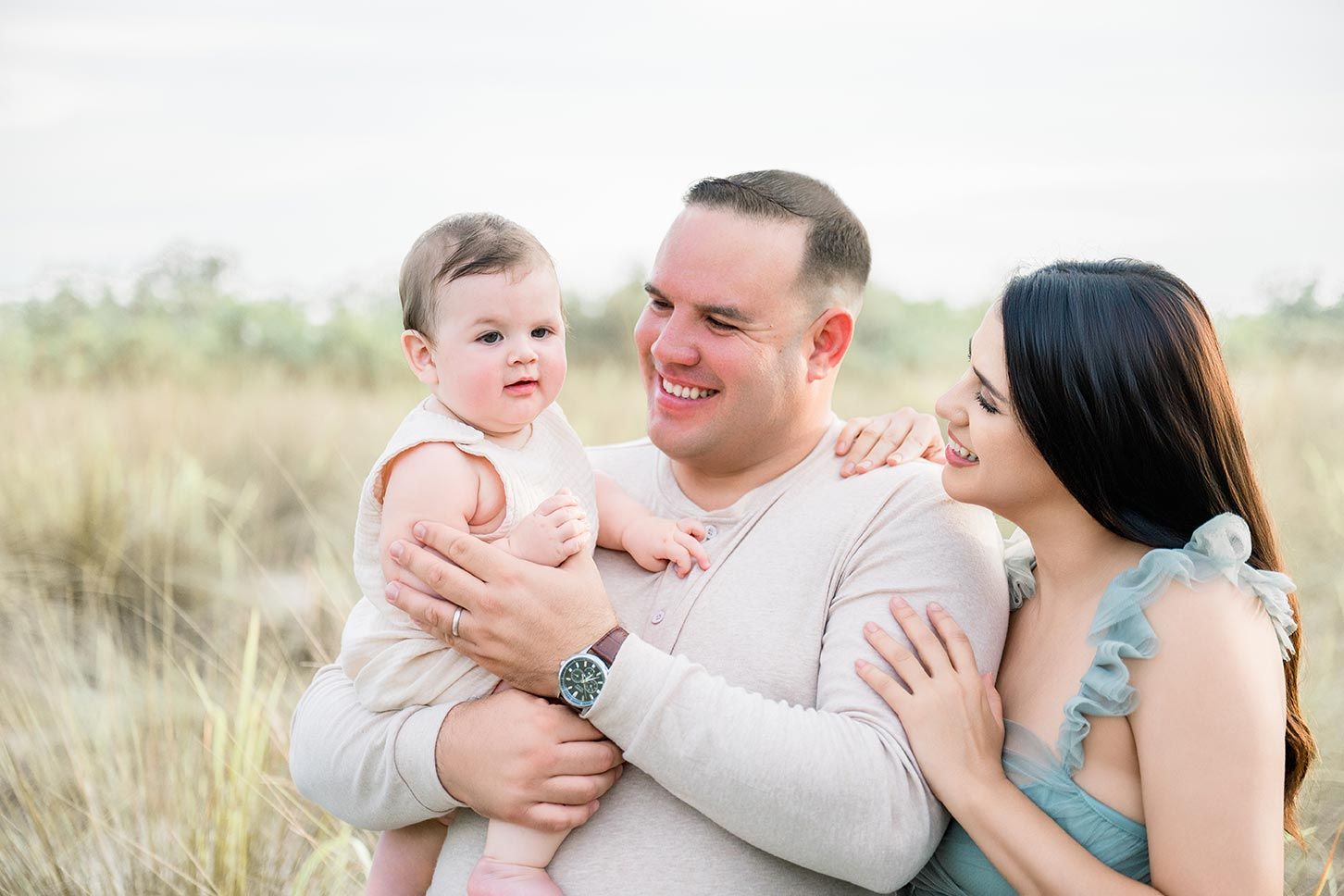 Happy family with smiling dad holding baby girl while mom looks on lovingly in a sunny outdoor field.