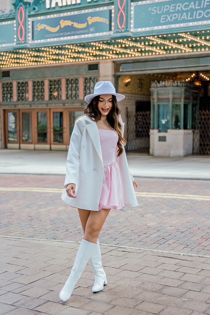 Woman in pink mini dress, white blazer, hat, and boots posing outside a theater.