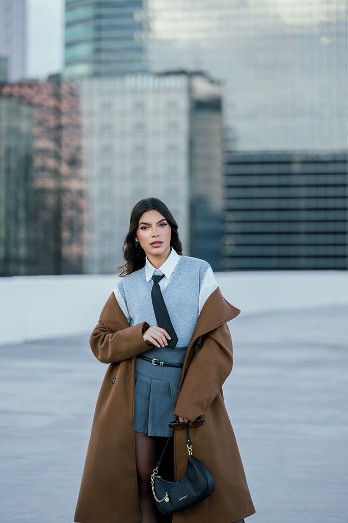 Woman in brown coat, gray sweater, tie, and pleated skirt holds black bag in urban setting.