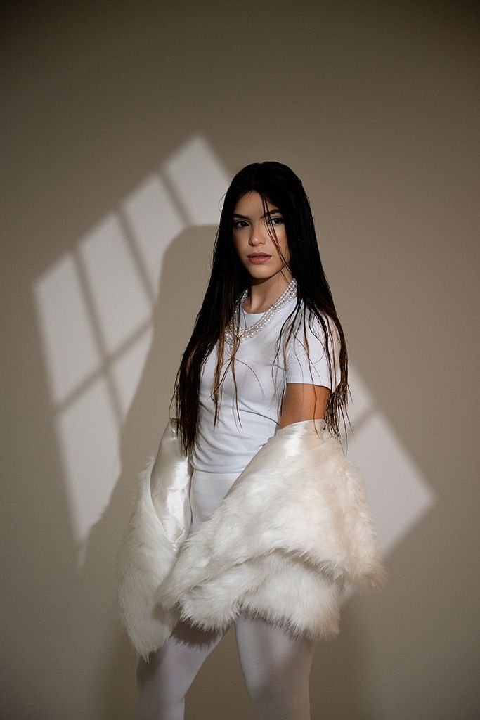 Young woman in white crop top, fur jacket, and leggings posing in dramatic window light against beige background.