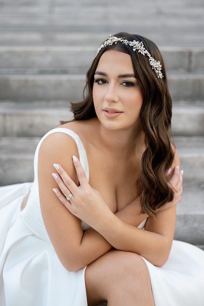 Elegant bride in white gown sits on stone steps, wearing a crystal hair vine, with soft waves and natural bridal makeup.