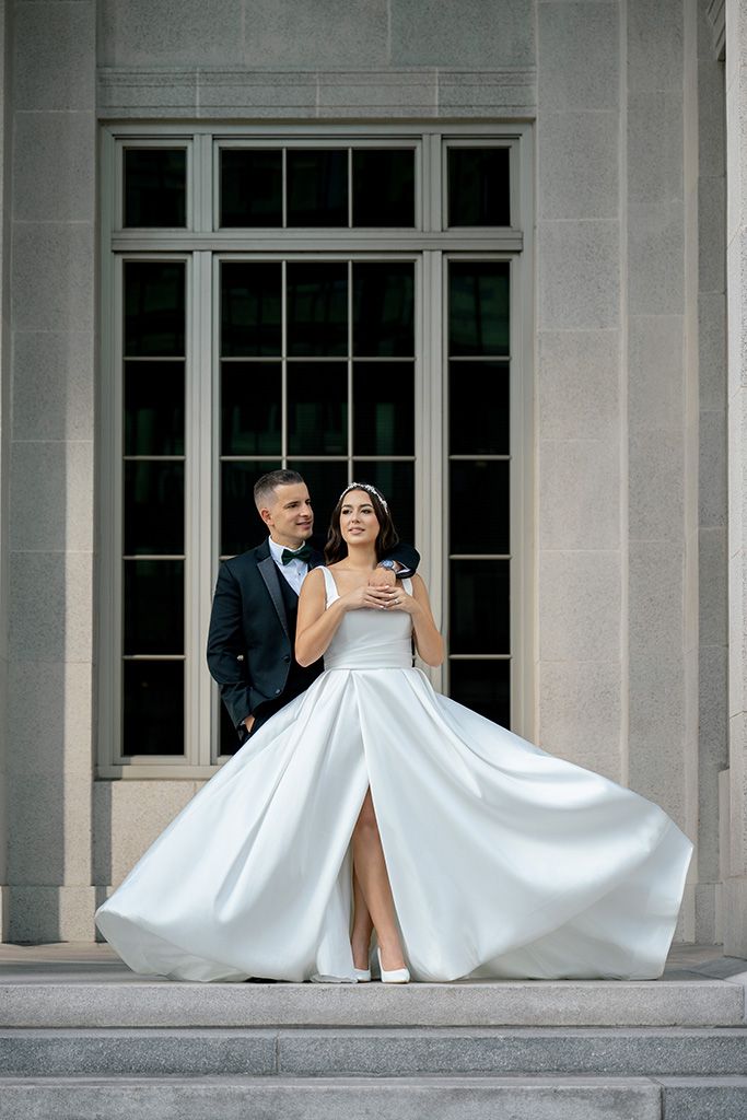 Elegant bride in flowing white gown with slit stands with groom in tuxedo on stone steps before grand building.