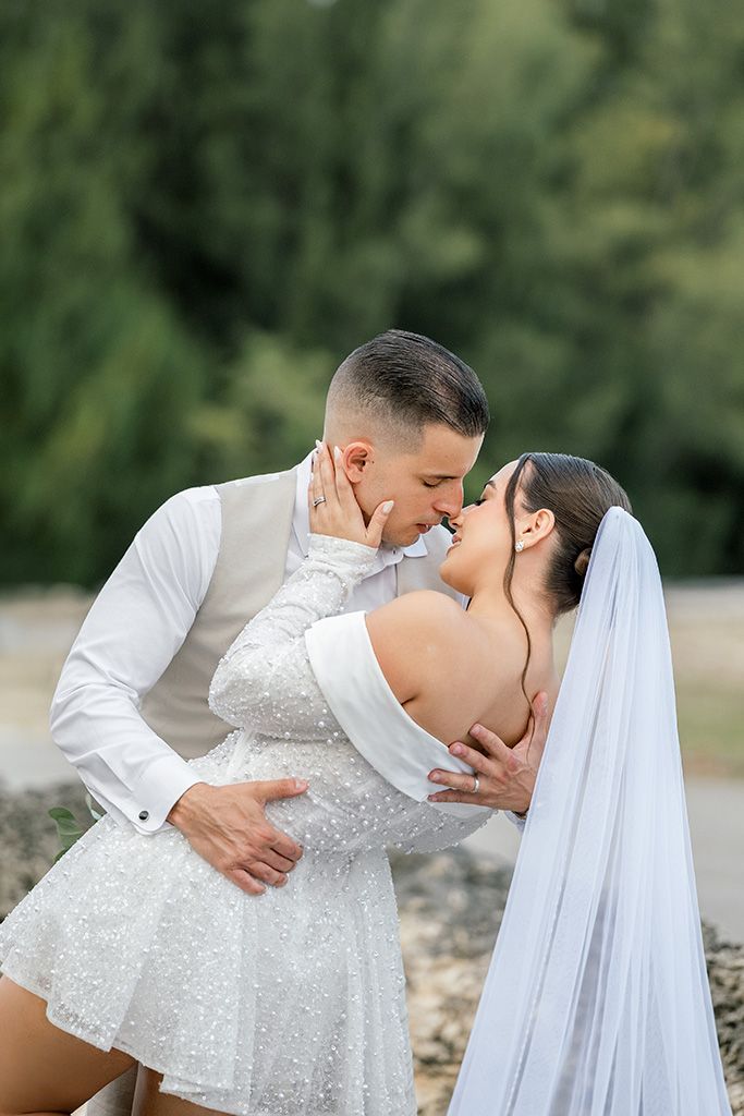 Newlyweds share a romantic kiss outdoors, bride in sparkly short wedding dress with veil, groom in white shirt and vest.