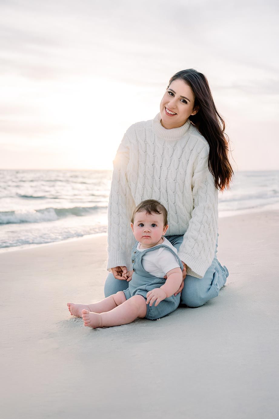 A relaxing sunset beach scene with someone wearing a cream cable knit sweater sitting next to a baby in blue.