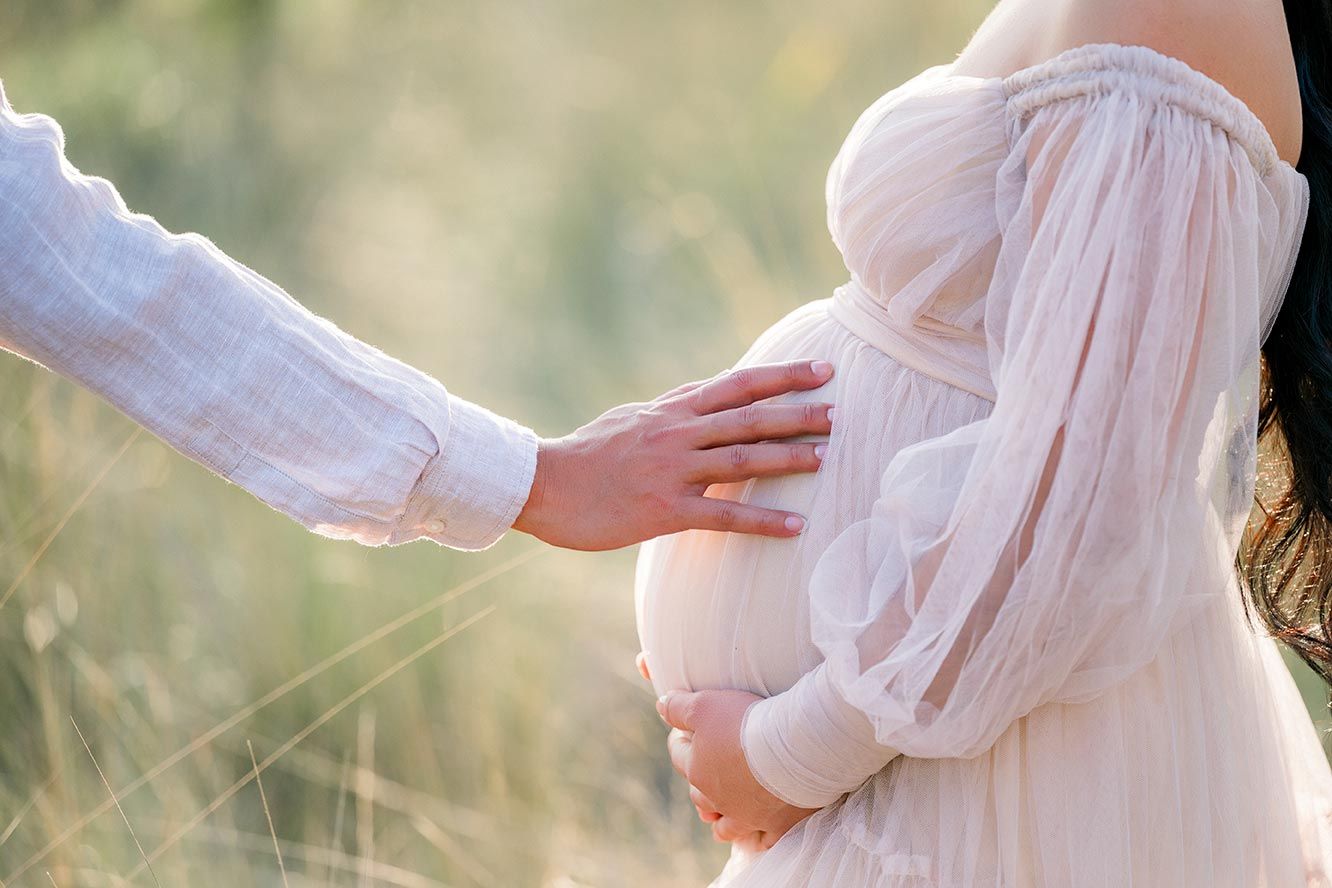 Maternity photoshoot set shows a woman in flowing white dress holding her baby bump outdoors in dreamy lighting.