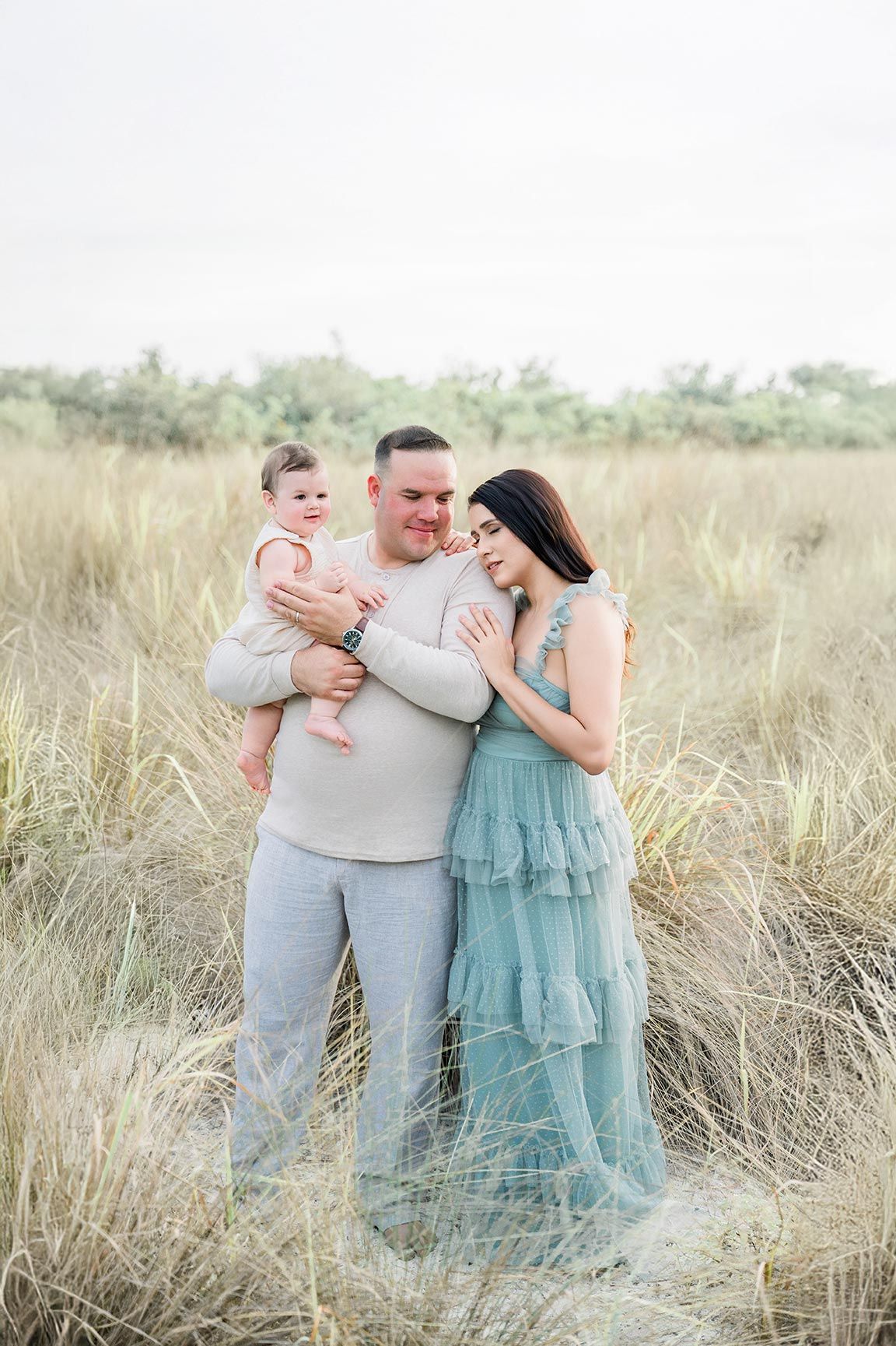 A loving family embraces in a sunlit field of tall grass, with the mother wearing a flowing turquoise dress.