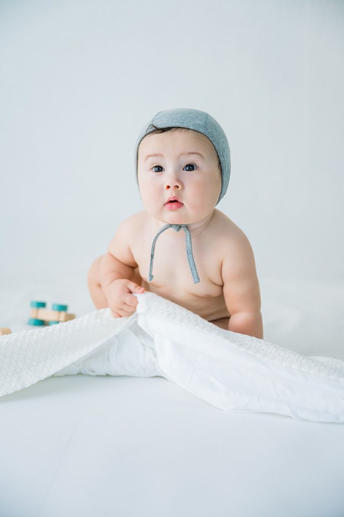Adorable baby in gray bonnet hat sitting on white blanket, shirtless with curious expression.
