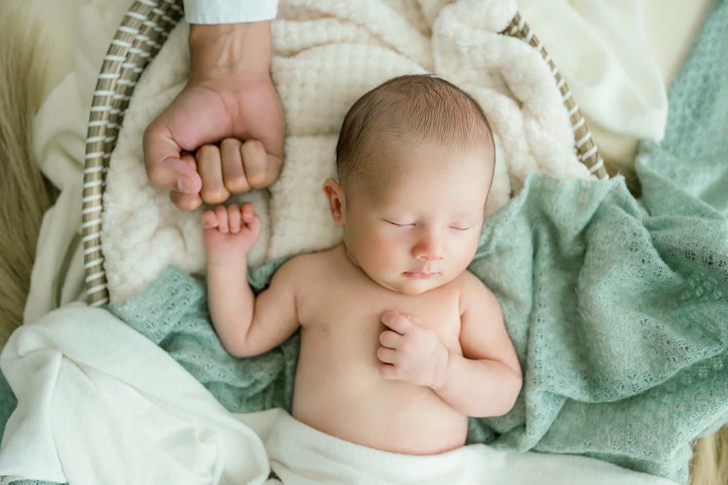 Peaceful newborn baby sleeping in a basket with a soft teal blanket, gently holding an adult's finger.