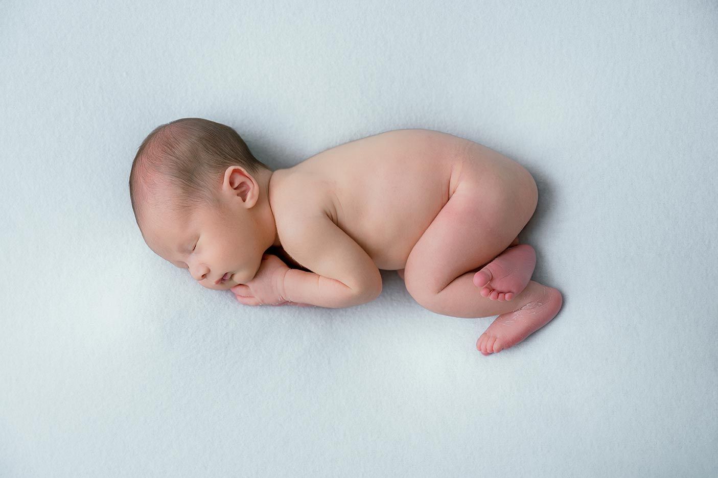 Peaceful newborn baby sleeping curled up on a soft white blanket in a classic posed newborn photography style.