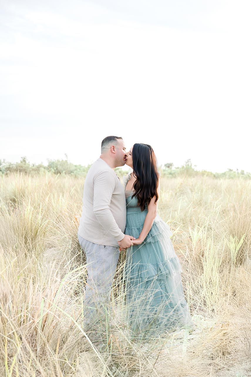 A couple shares a romantic kiss while holding hands in a sunlit golden grass field.