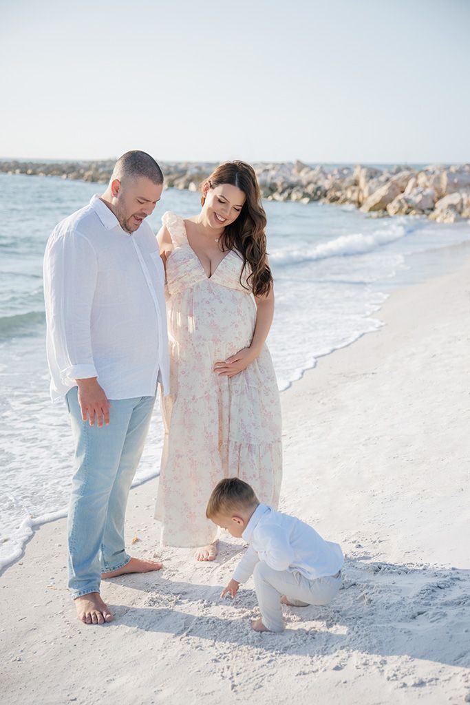 Pregnant couple in white attire watching their toddler play in the sand on a sunny Florida beach.
