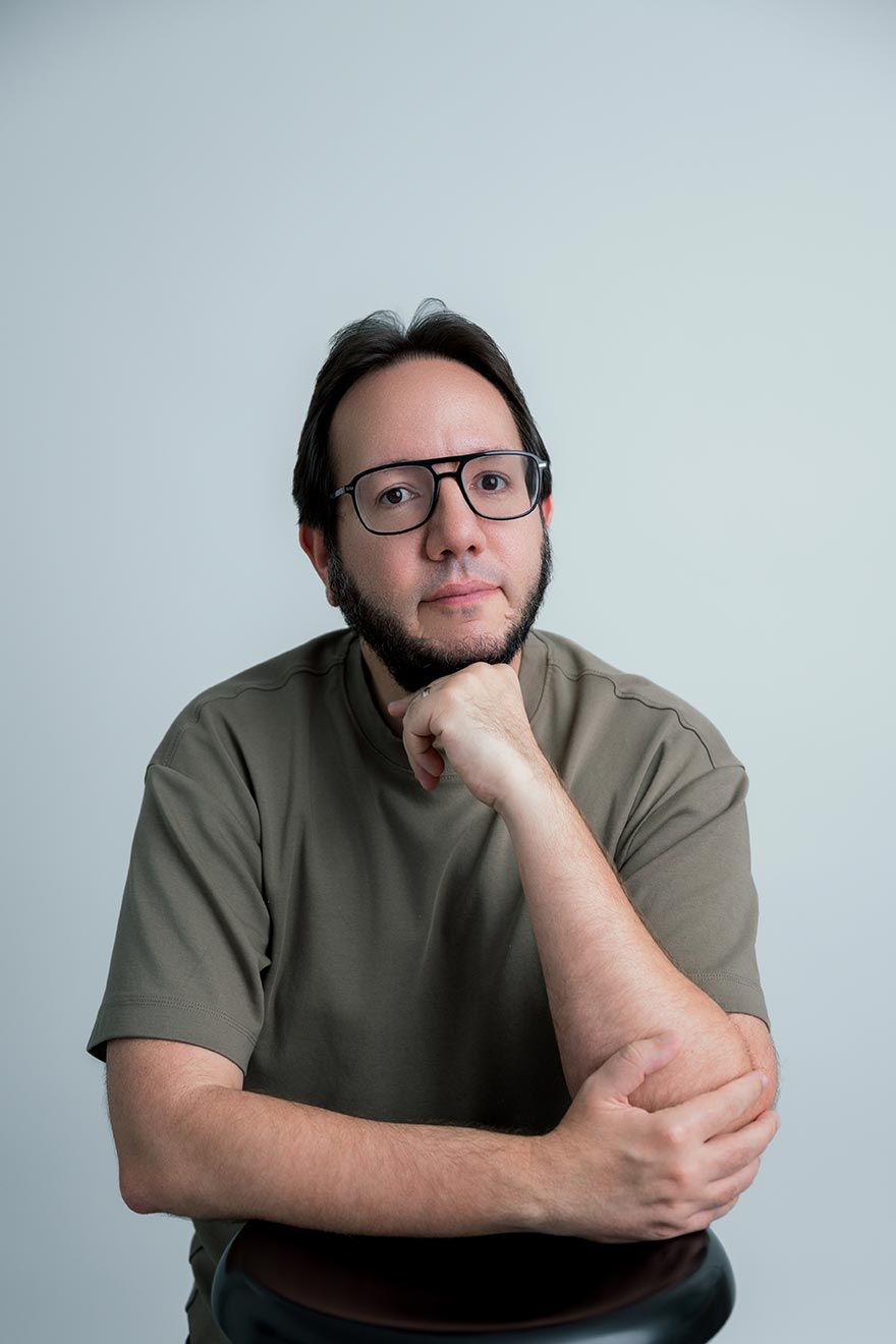 Portrait of someone in glasses and olive t-shirt with thoughtful pose against light gray background.