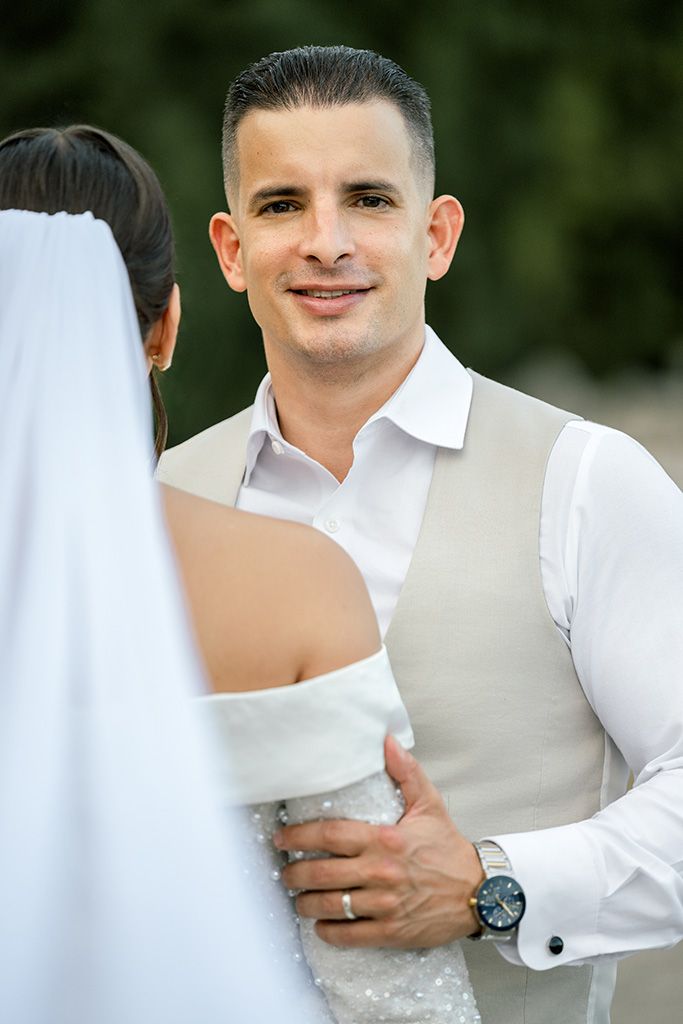 Smiling groom in white shirt and beige vest embraces bride outdoors during wedding celebration.