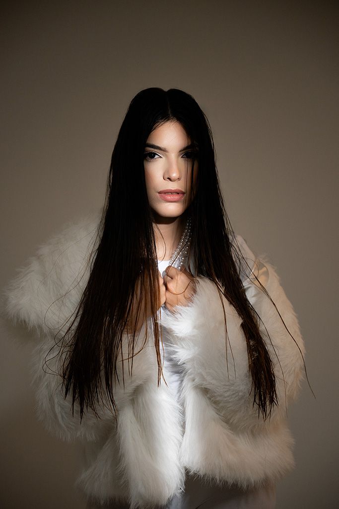 Young woman with long dark hair wearing a white fur coat and silver necklace posing against a neutral background.