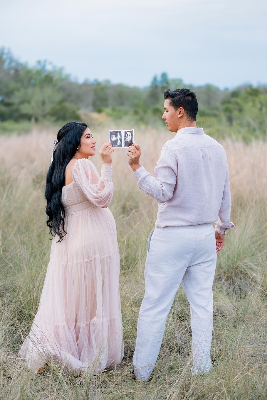 Couple holding ultrasound photos in a field during a maternity photoshoot, woman in pink gown.