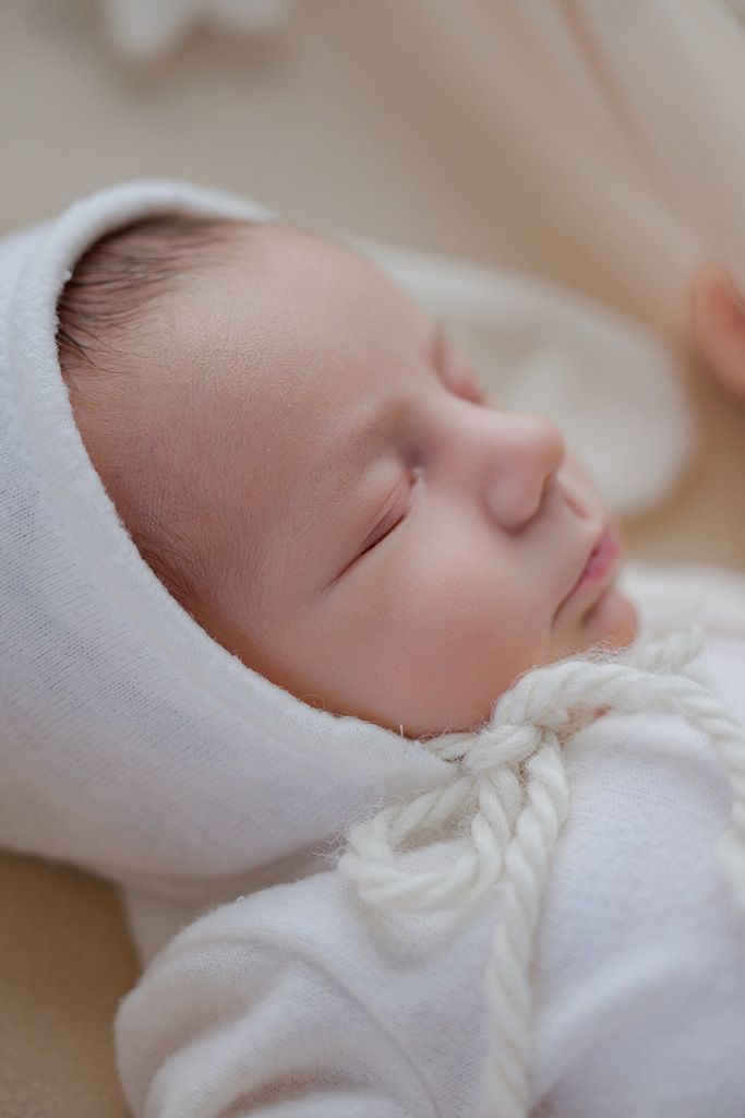 Close-up of a sleeping newborn baby wearing a white knit bonnet and soft white outfit with rope detail.