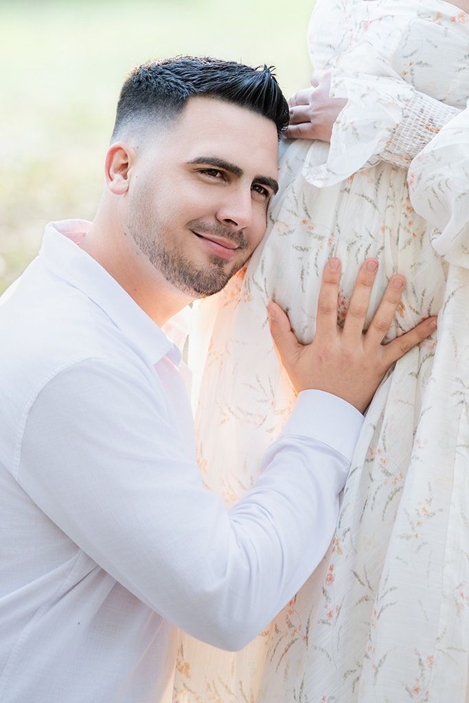 Man in white shirt tenderly rests his head against pregnant woman's lace maternity gown outdoors.