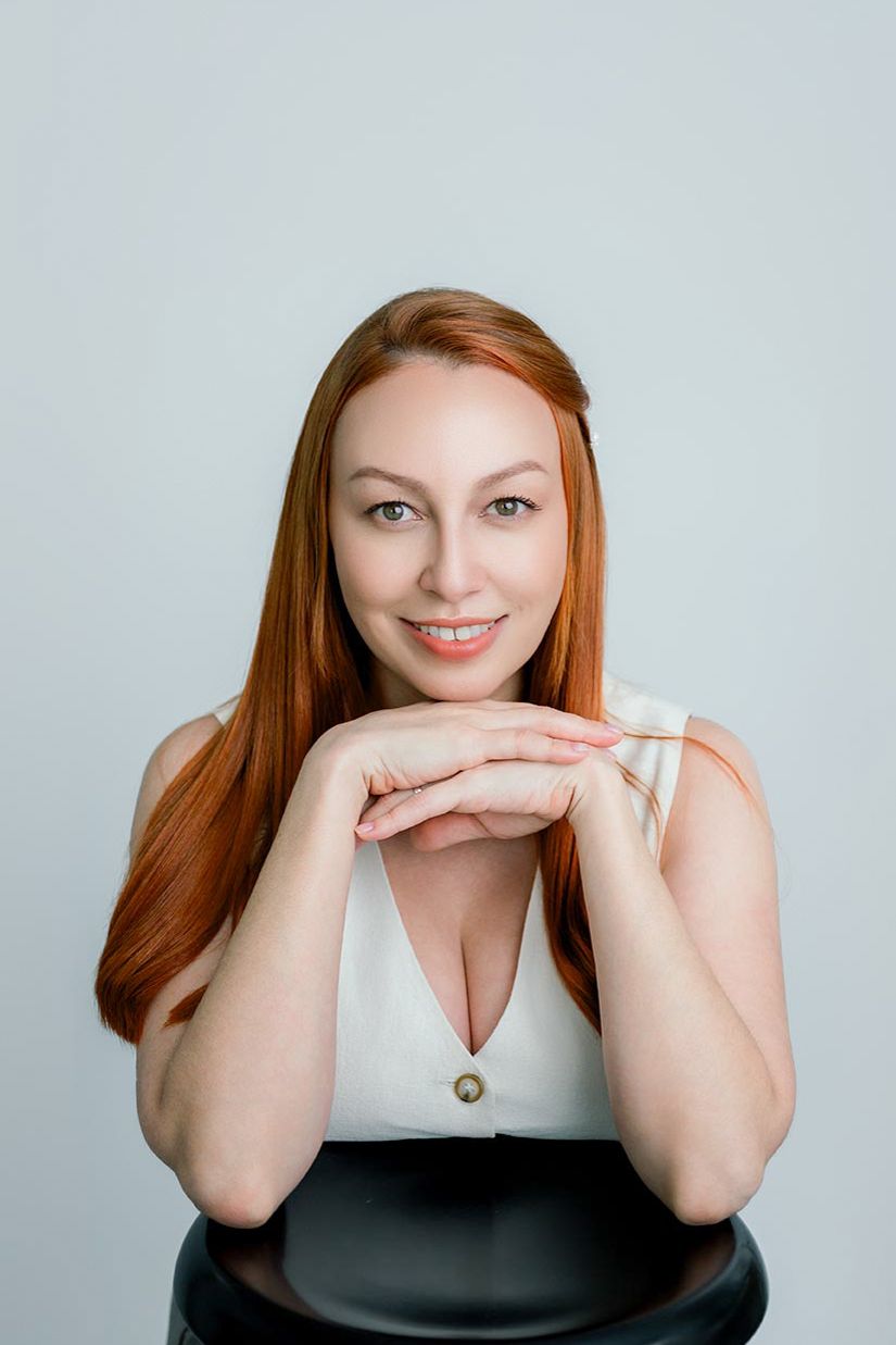 Portrait of someone with vibrant red hair wearing a white top, resting their chin on folded hands against a light background.