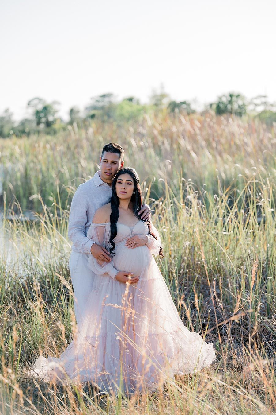 A romantic portrait of a couple embracing in a field of tall grass by the water's edge at golden hour.