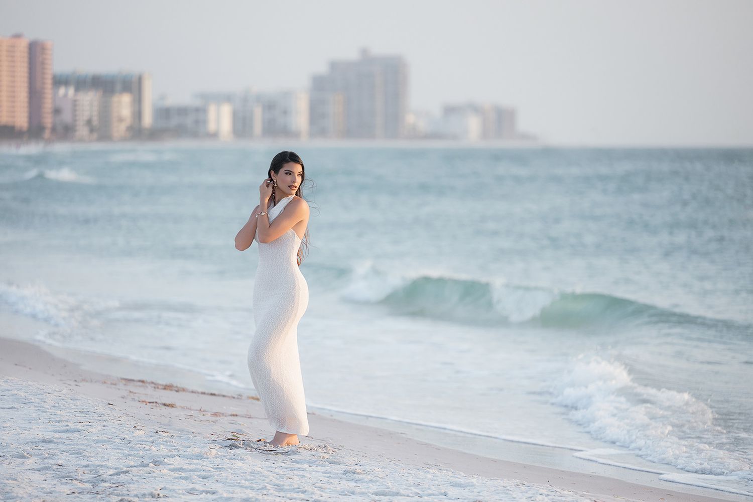 A woman in a white dress stands on a sandy beach with ocean waves and city skyline in the background.