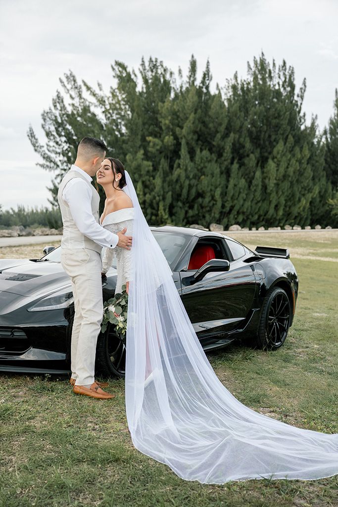 Bride in white gown with long veil and groom in cream suit pose beside a black Corvette sports car outdoors.