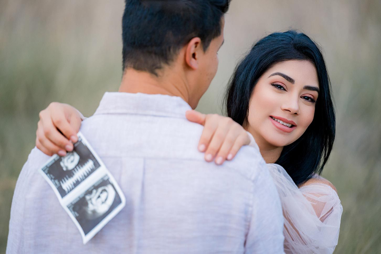 A couple poses outdoors while holding an ultrasound photo, announcing a pregnancy with joy.