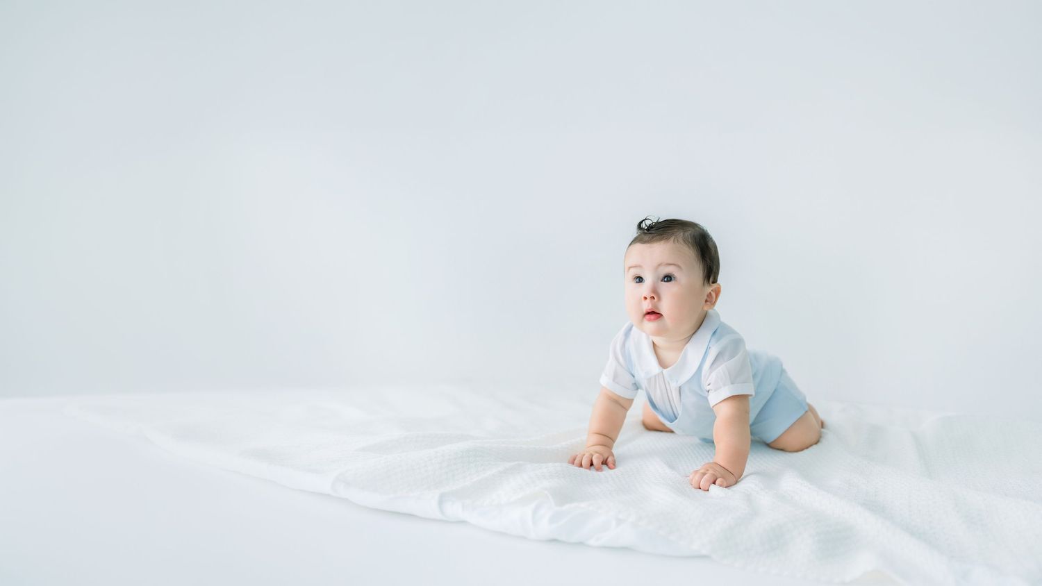 A cute Asian baby in a white onesie crawls on a white blanket against a clean white background.