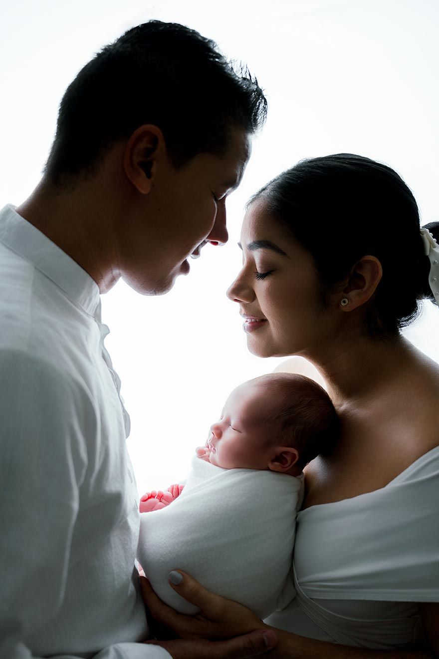 Silhouette of parents in white clothing lovingly holding their swaddled newborn baby together.