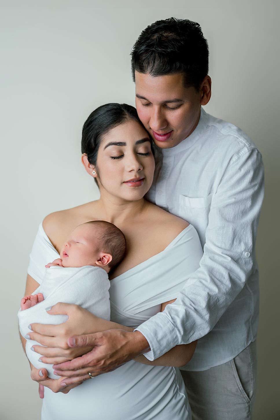 A young couple in white clothing tenderly holds their sleeping newborn baby wrapped in white cloth.