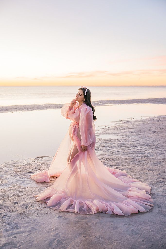 Woman in flowing pink gown poses gracefully on a serene beach at sunset, with sheer fabric trailing on sand.