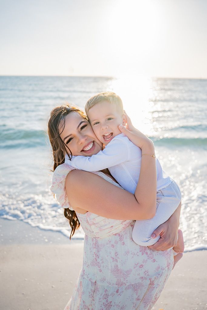 A smiling woman in a floral dress holds a laughing toddler on a sunny beach with turquoise waves behind them.