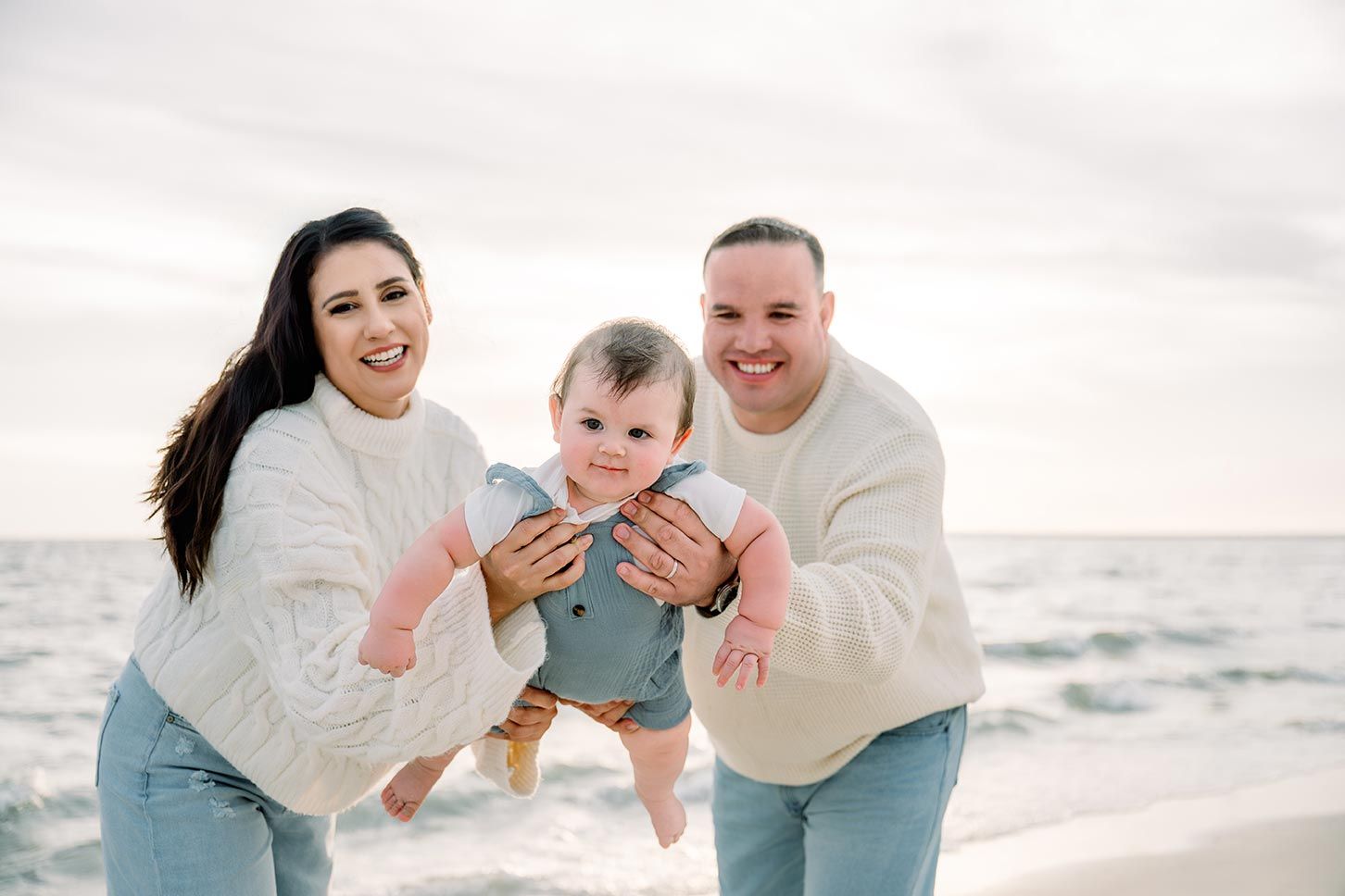 Happy family with baby at beach, parents holding infant over ocean waves during golden hour photo session.
